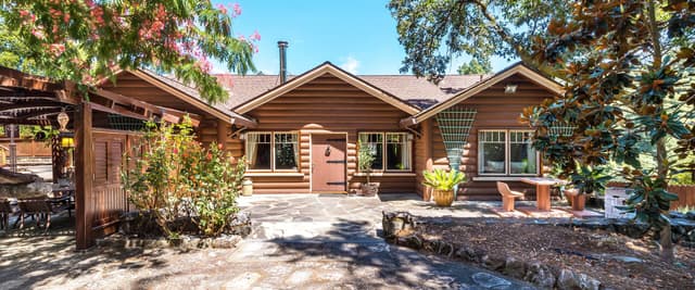Wooden lodge with turquoise swimming pool, lounge chairs, flowering trees, and thatched-roof gazebo surrounded by forest.